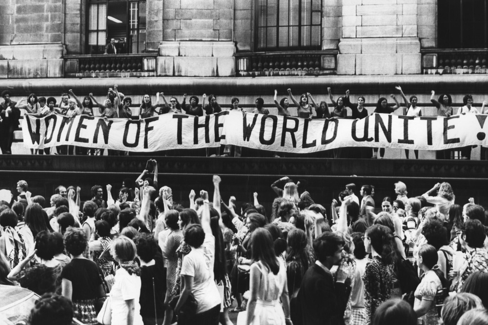 Black and white shot from a feminist demonstration, women with raised fists in the street and above them women on a balcony have posted alongside a banner that reads "women of the world unite!"