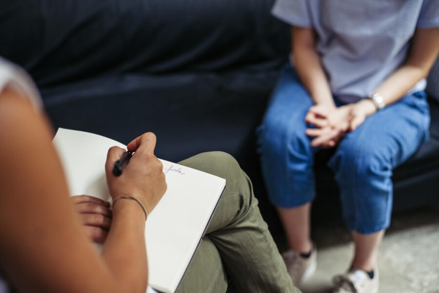 two women are sitting opposite each other, the first one has her hands on her knees and the second one is taking notes