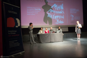 στη σκηνή δύο όρθιες γυναίκες συνομιλούν σε τέσσερα άτομα στο πάνελ, πίσω τους η προβολή της αφίσας «Μην προσπερνάς την έμφυλη βία» από προτζέκτορα