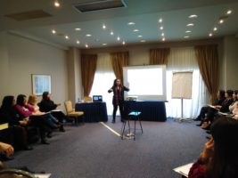 στιγμιότυπο από την επιμόρφωση, μια εκπαιδεύτρια δείχνει πάνω σε μία επιφάνεια παρουσίασης, οι συμμετέχουσες κάθονται σε ημικυκλική διάταξη