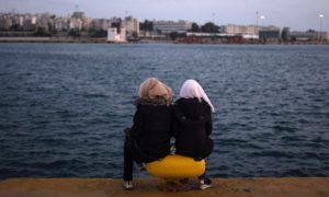 δύο προσφύγισσες κάθονται πάνω σε μια δέστρα λιμανιού και κοιτούν τη θάλασσα