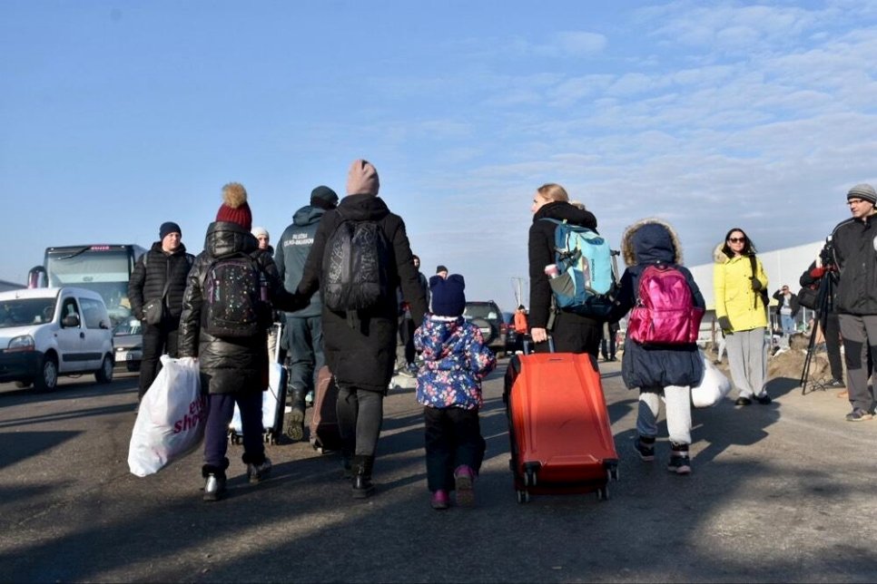 Φωτογραφία. Άτομα με βαλίτσες και παιδιά περπατάνε. Βλέπουμε τις πλάτες τους.