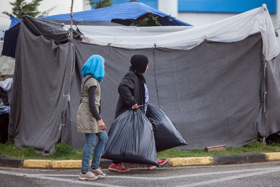 δύο γυναίκες μπροστά από μια σκηνή προσφυγικού καταυλισμού, η μία κρατά δύο γεμάτες μαύρες σακούλες σκουπιδιών στα χέρια της