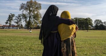 Φωτογραφία, δύο γυναικών πλάτυ που κοιτάνε ένα λιβάδι και φοράνε μια κίτρινη και μια μαυρη μαντίλα στο κεφάλι.