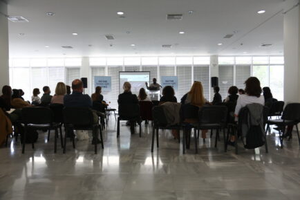 event shot, audience facing the panel where a speaker is standing, we see their backs