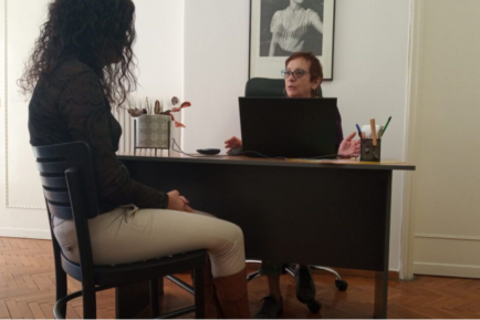 two women are talking, one sitting behind a computer desk and the other on the other side of the desk