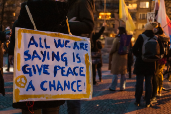 snapshot of an anti-war demonstration, a placard hanging around the neck of a protester reads "all we are saying is give peace a chance!"