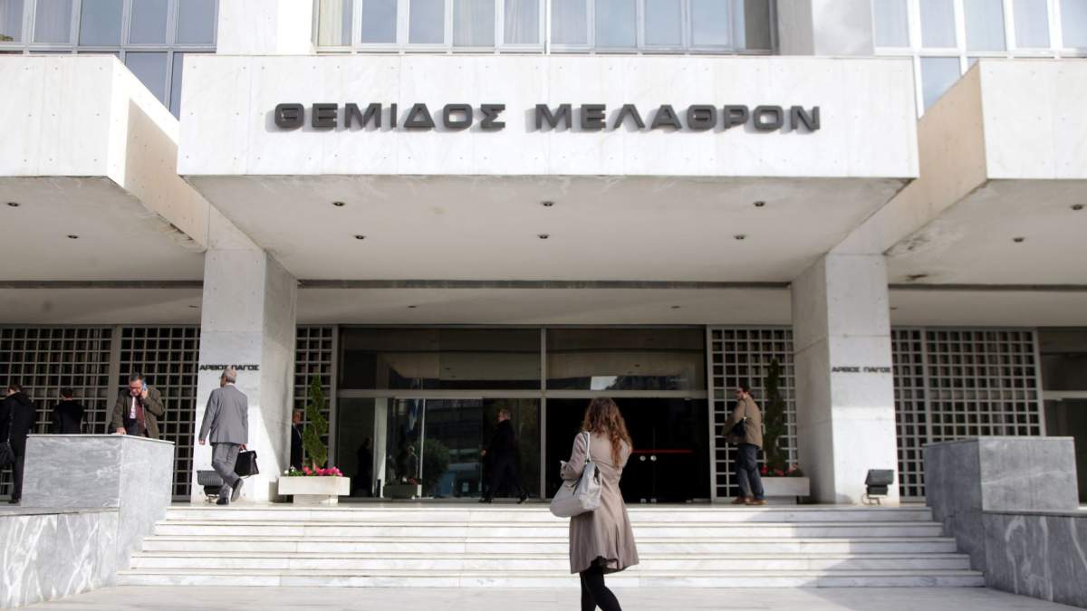 μια γυναίκα μπροστά από την είσοδο ενός κτιρίου που γράφει «Θέμιδος Μέλαθρον»