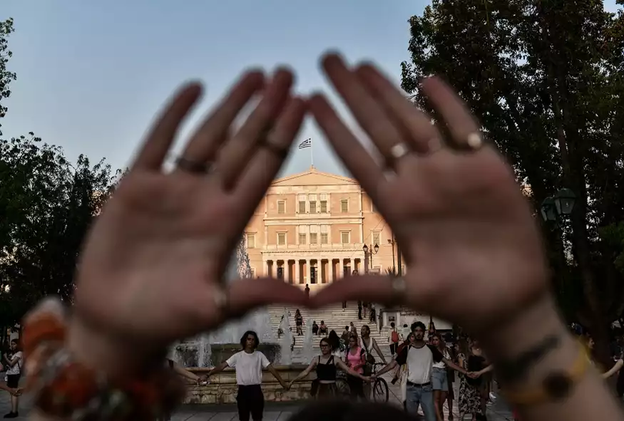 μια γυναίκα σχηματίζει ένα τρίγωνο με τις δύο παλάμες της, στο φόντο φαίνεται η Βουλή
