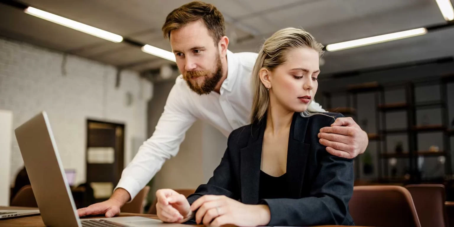 μια εργαζόμενη σε γραφείο κοιτά ενοχλημένη έναν άνδρα να στέκεται από πάνω της και να την ακουμπά στον ώμο