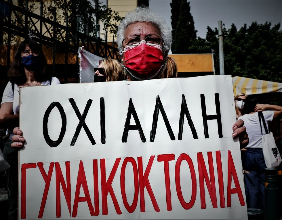 μια διαδηλώτρια με κόκκινη μάσκα προστασίας κρατά ένα λευκό χαρτόνι που γράφει με μαύρα και κόκκινα γράμματα «όχι άλλη γυναικοκτονία»