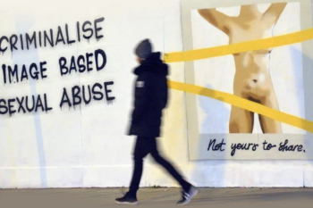 ένας άνδρας περνά μπροστά από μία αφίσα που δείχνει ένα γυμνό σώμα με κάλυψη στο στήθος και τα γεννητικά όργανα η λεζάντα γράφει «not yours to share» και «criminalise image based sexual abuse»