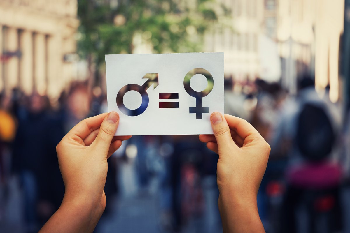 μια γυναίκα κρατά ένα χαρτί που απεικονίζει το σύμβολο της ισότητας και εκατέρωθεν του το αρσενικό και θηλυκό σύμβολο
