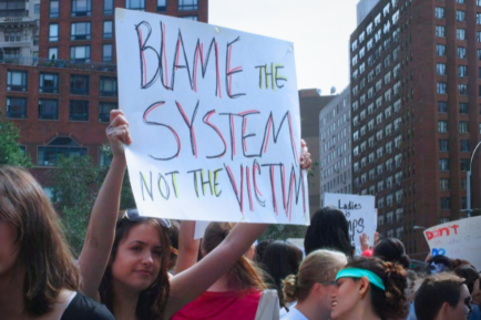 φωτογραφία με γυναίκα που κρατάει πλακάτ "blame the system not the victim" που πλαισιώνει άρθρο για μια καταγγέλλουσα που βρέθηκε κατηγορούμενη