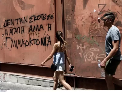 μια γυναίκα βαδίζει δίπλα σε έναν τοίχο που γράφει «Δεν τη σκότωσε η αγάπη αλλά η γυναικοκτονία»