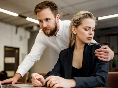 μια εργαζόμενη σε γραφείο κοιτά ενοχλημένη έναν άνδρα να στέκεται από πάνω της και να την ακουμπά στον ώμο