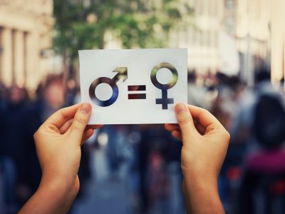 μια γυναίκα κρατά ένα χαρτί που απεικονίζει το σύμβολο της ισότητας και εκατέρωθεν του το αρσενικό και θηλυκό σύμβολο