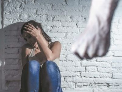 μια γυναίκα κάθεται με την πλάτη στον τοίχο, ένας άνδρας με σφιγμένη γροθιά στέκεται μπροστά της